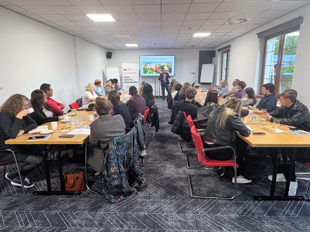 Uczestnicy szkolenia siedzą przy dwóch podłużnych stołach. W tel, przy ekranie, stoi prowadzący. Po lewej obok ekranu rollup z tytułem projektu.
