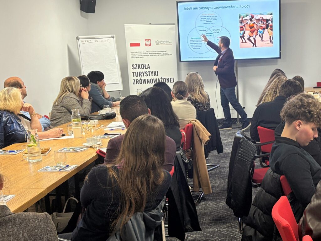 Grupa uczestników szkolenia „Szkoła Turystyki Zrównoważonej” siedzi przy stołach konferencyjnych i słucha wykładu. Trener stojący przy ekranie wskazuje schemat dotyczący turystyki zrównoważonej, obok którego widoczna jest fotografia tańczącej grupy ludzi. W tle roll-up z logo Ministerstwa Sportu i Turystyki oraz tytułem projektu.